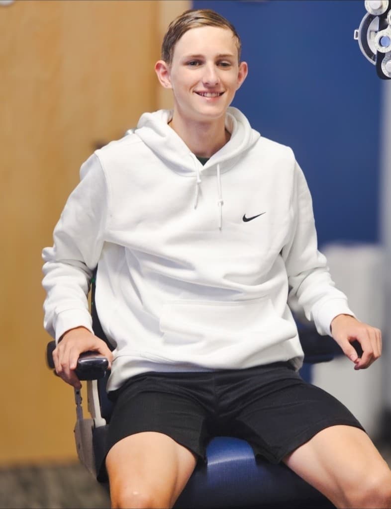 Branson smiling in a blue exam chair at Brazos Eye Surgery, wearing a white Nike hoodie during a visit to the clinic
